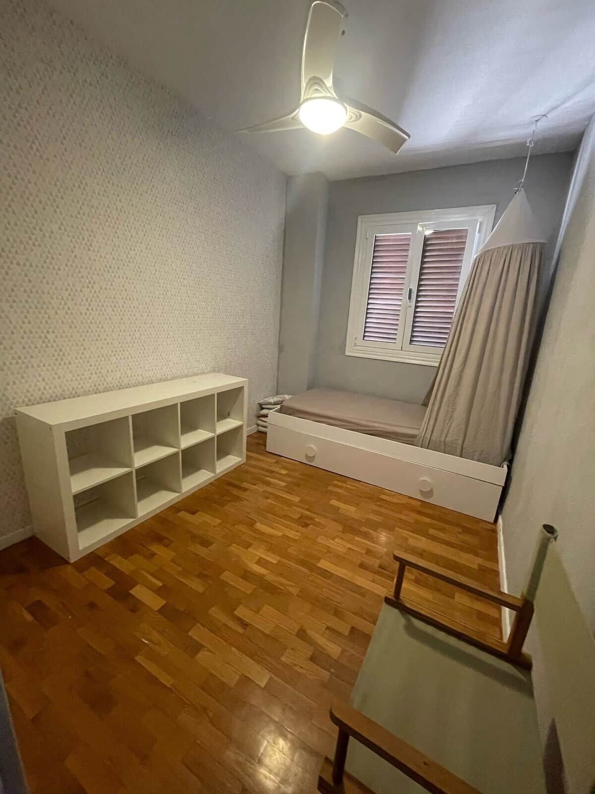 A room with a wooden floor, a white built-in bench, a ceiling fan, a window with blinds, and a bed with a headboard.