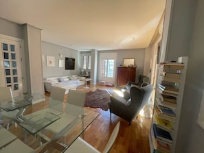 A spacious living area with a glass dining table, a sofa, a TV, and a bookshelf.