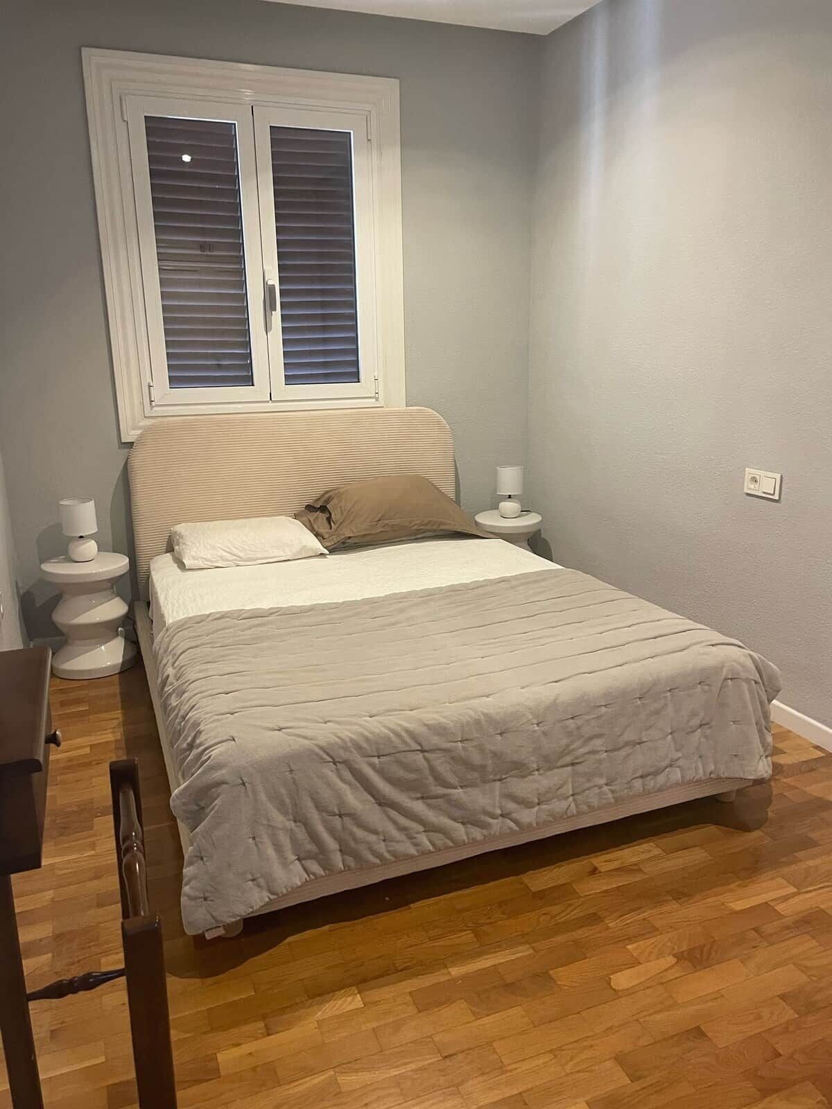 Ein Schlafzimmer mit Bett, Fenster mit Jalousien, Nachttisch mit Lampe und Holzboden.