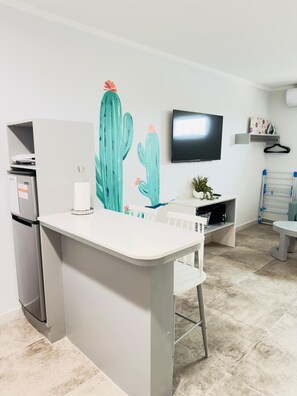 A modern kitchen with a white countertop, a refrigerator, a flat-screen TV mounted on the wall, and a small potted plant on the counter.