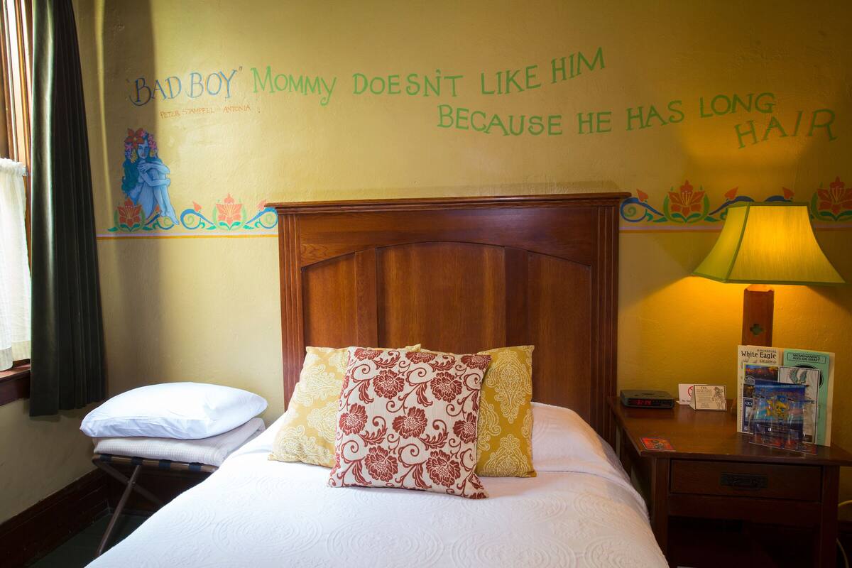 A hotel room with a bed, pillows, a nightstand with a lamp, and a wall with a humorous quote.