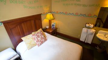 A hotel room with a bed, a nightstand with a lamp, a wall with humorous text, a bathroom with a sink and towels.