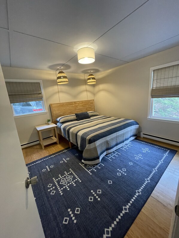 Ein Schlafzimmer mit Bett, Nachttischen, einem Fenster mit Jalousien, einem gemusterten Teppich und Pendelleuchten.