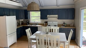 A modern kitchen with dark blue cabinets, a white refrigerator, a stove, and a dining table with white chairs. - Into the Woods (Southwest Harbor)