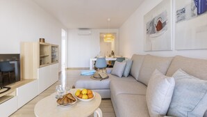 A modern living room with a sofa, a dining table with a bowl of fruit and pastries, and a kitchen area in the background. - Golden Paradise Mar (Javea / Xàbia)