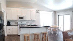 A modern kitchen with white cabinetry, a black countertop, and a built-in microwave. There are three wooden stools at the island, and a dining table with chairs in the background. - Affordable Gathering Place-90 minutes to Yellowstone (Idaho Falls)
