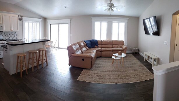 A spacious living area with a large L-shaped sectional sofa, a kitchen with white cabinets, a dining area with stools, and a flat-screen TV mounted on the wall. - Affordable Gathering Place-90 minutes to Yellowstone (Idaho Falls)