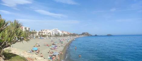 Une plage où des gens se prélassent au soleil et se baignent, avec des bâtiments en arrière-plan et un ciel dégagé.