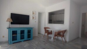 Una habitación con un televisor de pantalla plana montado en la pared, un mueble azul con puertas de vidrio, dos sillas de mimbre alrededor de una pequeña mesa blanca y una ventana con vistas a una zona de cocina.