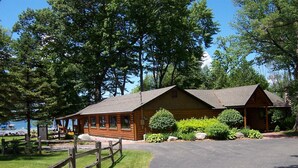 A rustic log cabin with a porch, surrounded by lush greenery and a clear sky.