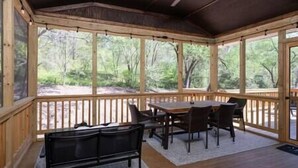 Un área de estar al aire libre techada, con mesa y sillas, barandas de madera y vistas al bosque circundante.