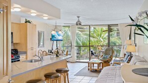A modern kitchen with a bar area, a dining space with a view of palm trees, and a cozy seating area with a large bed. - Maui Sunset A303 - Great large 1 bedroom close to ocean (Kihei)