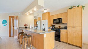 A modern kitchen with wooden cabinets, a central island with stools, a microwave, stove, and a refrigerator. - Maui Sunset A303 - Great large 1 bedroom close to ocean (Kihei)