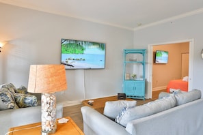 Ein Wohnzimmer mit Fernseher, der eine Strandansicht zeigt, einer blauen Kommode mit dem Schriftzug 'Beach' und einem grauen Sofa.