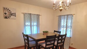 A dining area with a wooden table and four chairs, a chandelier, and two windows with patterned curtains.
