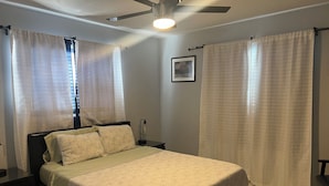 Un dormitorio con cama, mesitas de noche, ventilador de techo y cortinas.