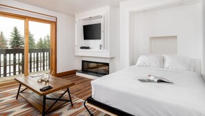 A modern hotel room with a large bed, a flat-screen TV mounted on a white wall, a fireplace, and a balcony with a wooden deck.