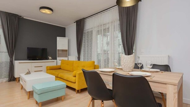 A modern living room with a yellow sofa, a dining table with chairs, a white cabinet, and a television.