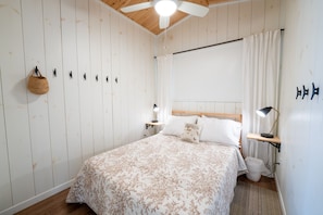 A bedroom with a bed, a ceiling fan, a bedside table with a lamp, a hanging basket, and a wall-mounted shelf.