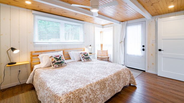 A bedroom with a wooden ceiling, a large bed with floral bedding, a nightstand with a lamp, and a window with blinds. - Family Accessible Lakehouse (Westport)