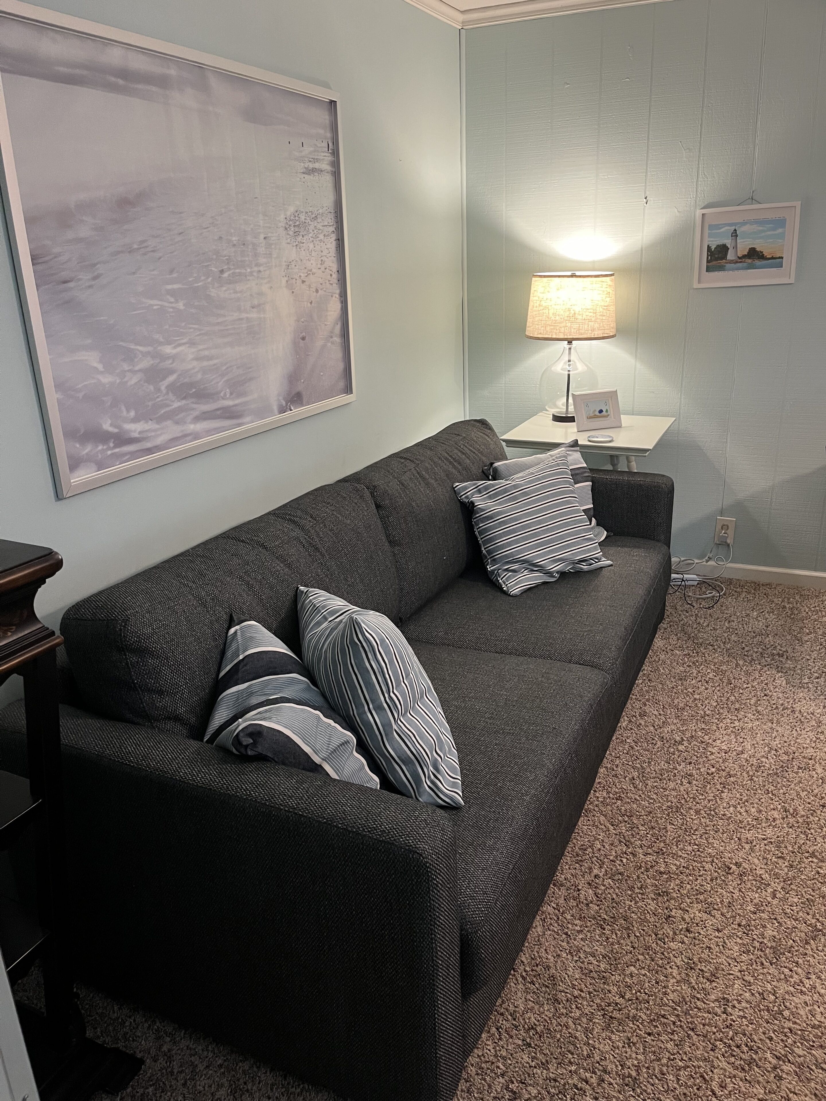 Un salon avec un canapé gris foncé, des coussins à rayures, un grand tableau abstrait, une table avec une lampe et un tableau encadré accroché au mur.
