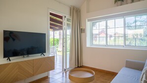 Una sala de estar con televisor de pantalla plana, un sofá, una mesa redonda de madera y una ventana con vistas a zonas verdes y edificios.