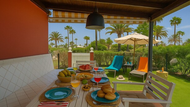 Un área de comedor al aire libre con una mesa lista para una comida, rodeada de sillas y sombrillas, con vista a palmeras y una cerca de piquetes blancos.