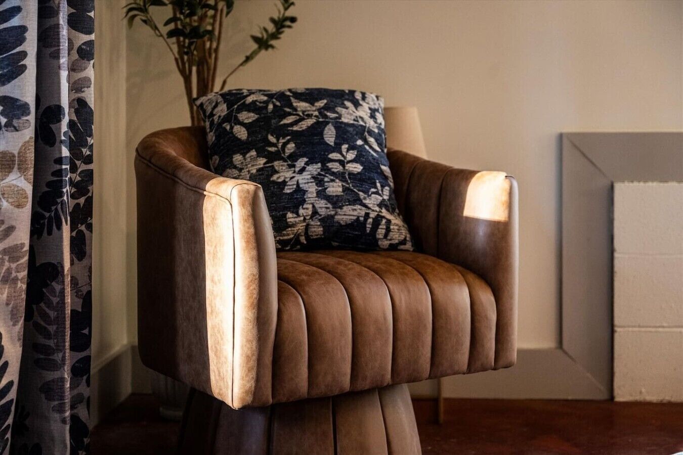 Un sillón de cuero marrón con un cojín floral, ubicado en una habitación con una cortina y una planta al fondo.