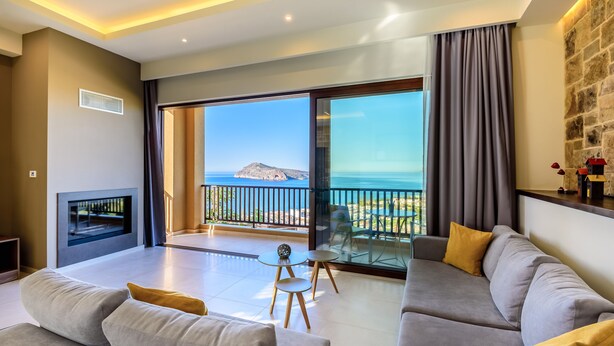 A modern living room with a sofa, a small table, and a fireplace. There is a balcony with a view of the sea and a mountain. - Sunset Villa - Panorama Sea View Villa (Platanias)