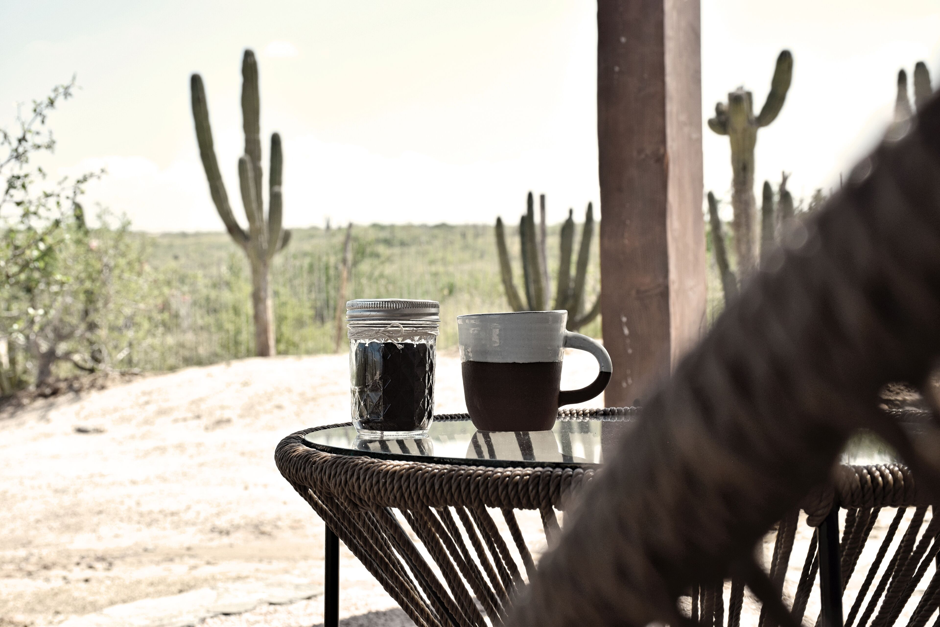 A table with two cups and a jar, set against a backdrop of cacti and a dirt path.