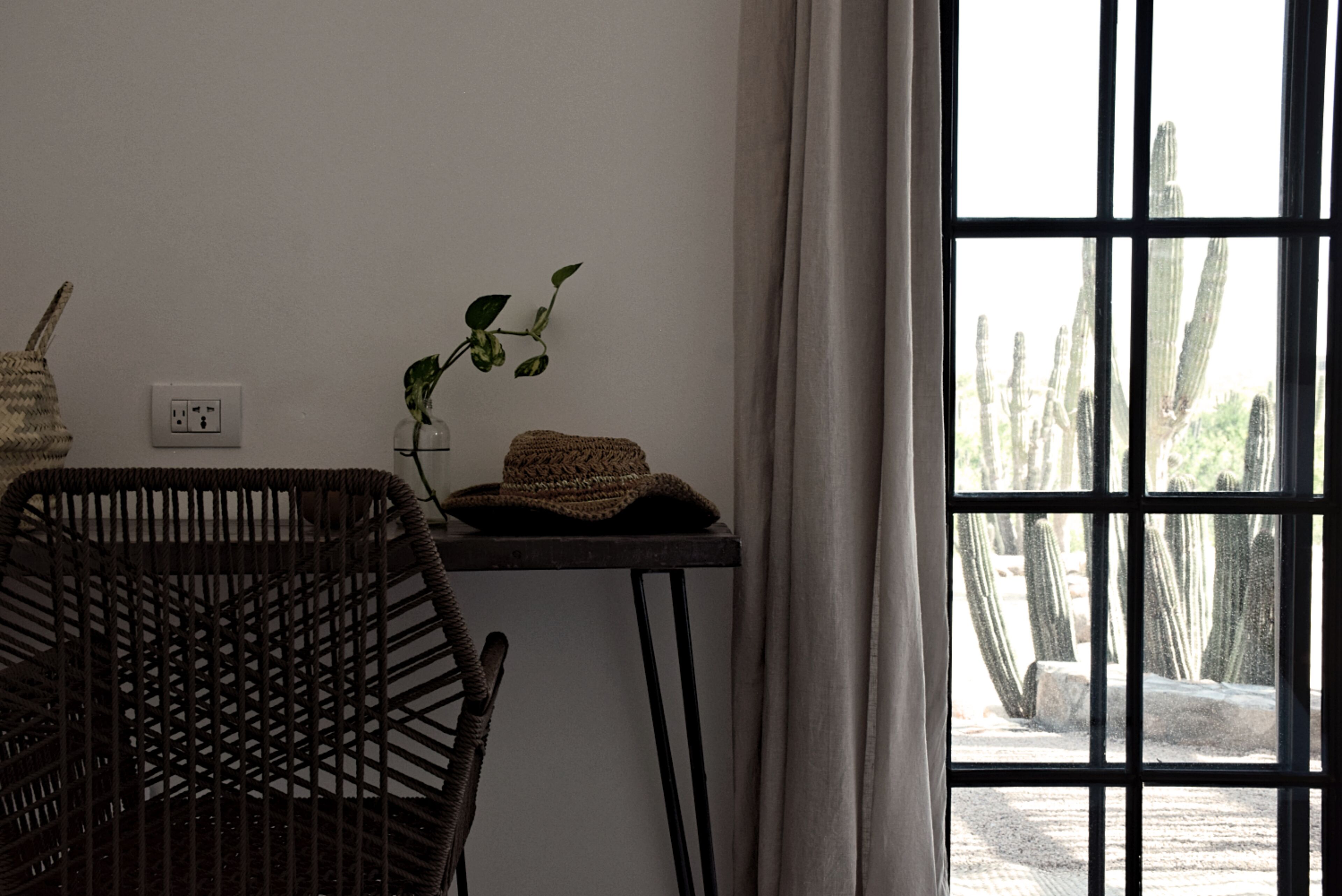 Ein Zimmer mit einem Korbstuhl, einem kleinen Tisch mit Pflanze und gewebtem Korb sowie einem Fenster mit Blick auf Kakteen.