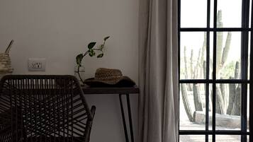 Ein Zimmer mit einem Korbstuhl, einem kleinen Tisch mit Pflanze und gewebtem Korb sowie einem Fenster mit Blick auf Kakteen.
