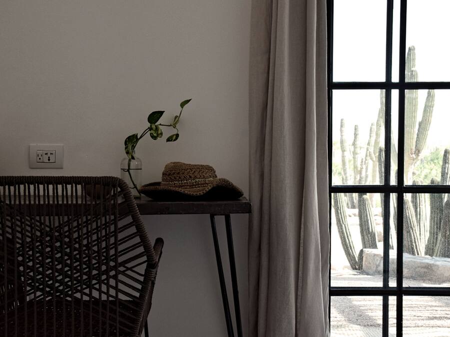 Ein Zimmer mit einem Korbstuhl, einem kleinen Tisch mit Pflanze und gewebtem Korb sowie einem Fenster mit Blick auf Kakteen.