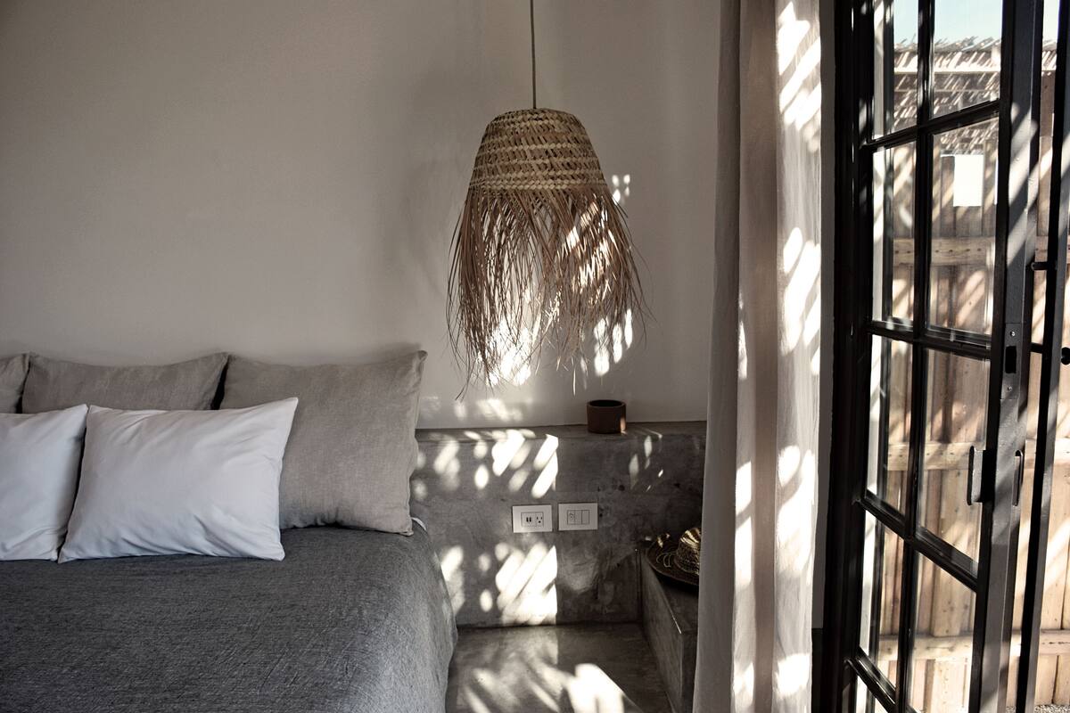Un dormitorio con cama, almohadas, una lámpara de fibra y vistas a través de una ventana.