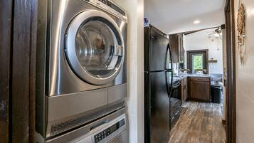 A laundry area with a front-loading washer and dryer, a black refrigerator, and a kitchen with wooden flooring and a window.