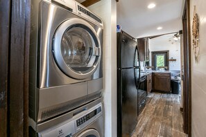 Zona de lavandería con lavadora y secadora de carga frontal, un refrigerador negro y una cocina con piso de madera y ventanal.