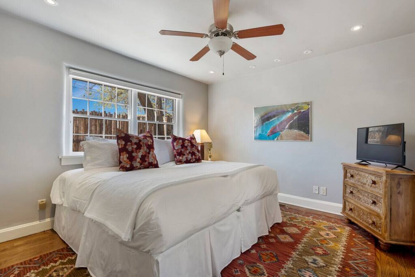 A bedroom with a large bed, a ceiling fan, a window with blinds, a TV on a wooden cabinet, and a rug.