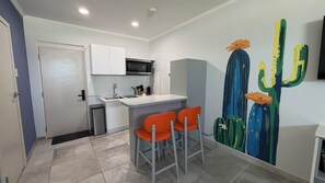 Une cuisine compacte avec des meubles blancs, un four à micro-ondes et un évier. On y trouve deux chaises orange et une fresque murale représentant un cactus.