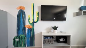 Une pièce décorée d’un tableau représentant un cactus, avec une télévision, une petite plante et un coffre-fort posé sur une console blanche.