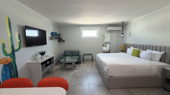 A hotel room with a bed, a television, a desk, a chair, a small table, and a cactus painting.