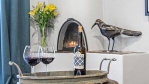 Una bandeja con dos copas de vino y una botella sobre una mesa, un florero con flores amarillas y una chimenea con una escultura de un pájaro.