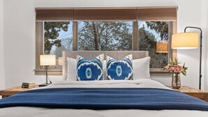 Un dormitorio con cama, dos almohadas, una mesita de noche, una lámpara y una ventana con vistas a árboles.