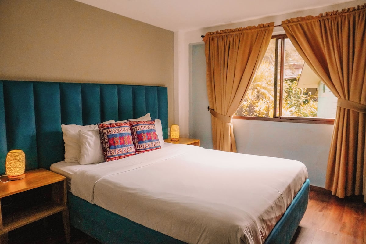 A neatly made bed with a teal headboard, white bedding, and patterned pillows. There are two bedside lamps on wooden nightstands. The room has a window with beige curtains and a view of greenery outside.