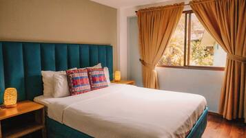 Una cama bien hecha con cabecera color turquesa, ropa de cama blanca y almohadas estampadas. Hay dos lámparas de noche sobre mesitas de madera. La habitación tiene una ventana con cortinas color beige y vistas a la vegetación exterior.