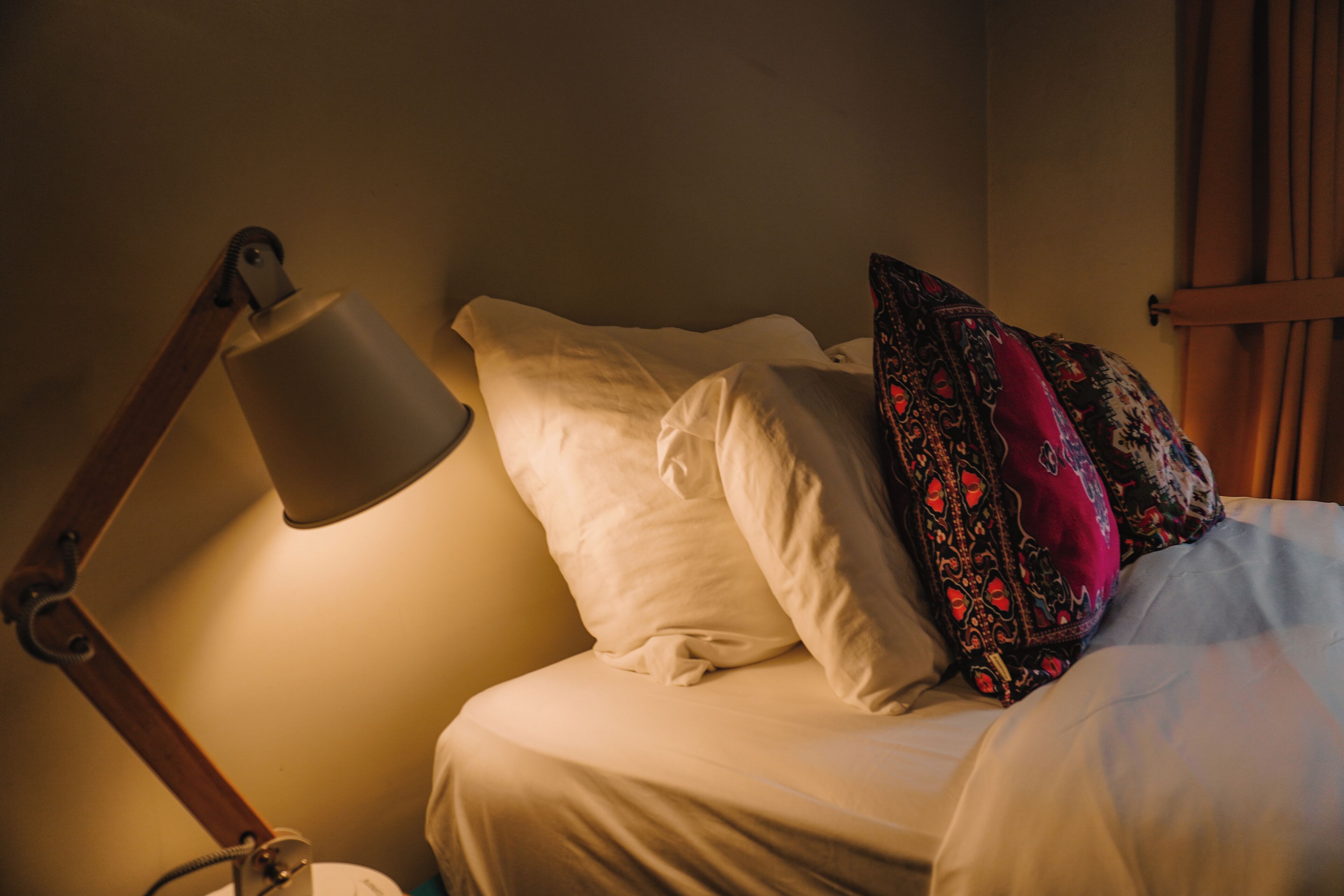 Una cama con ropa de cama blanca y una única almohada roja con estampado. Una lámpara de escritorio está colocada a la izquierda de la cama.