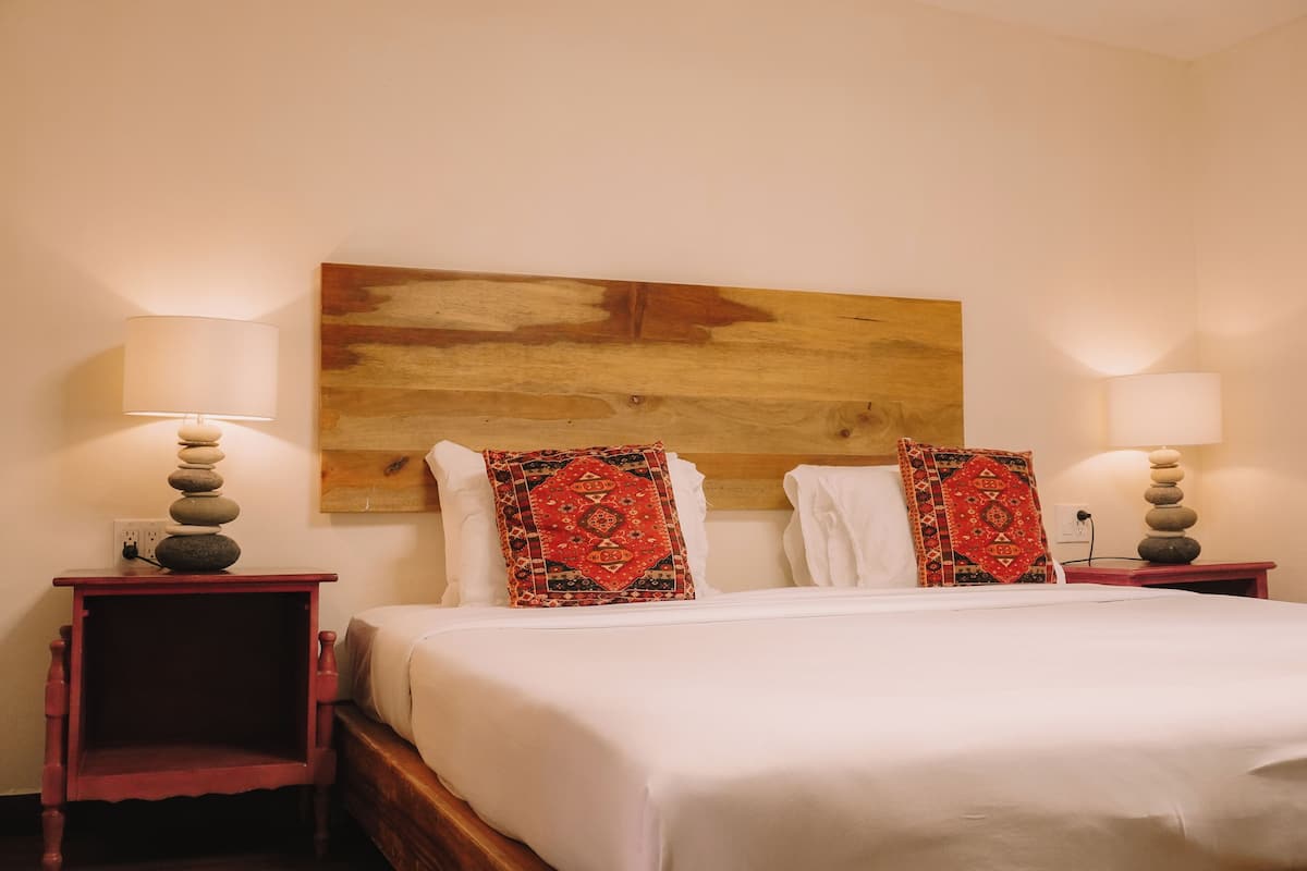 A neatly made bed with white linens, two decorative pillows, and a wooden headboard. There are two bedside tables with lamps and a wall-mounted artwork above the bed.
