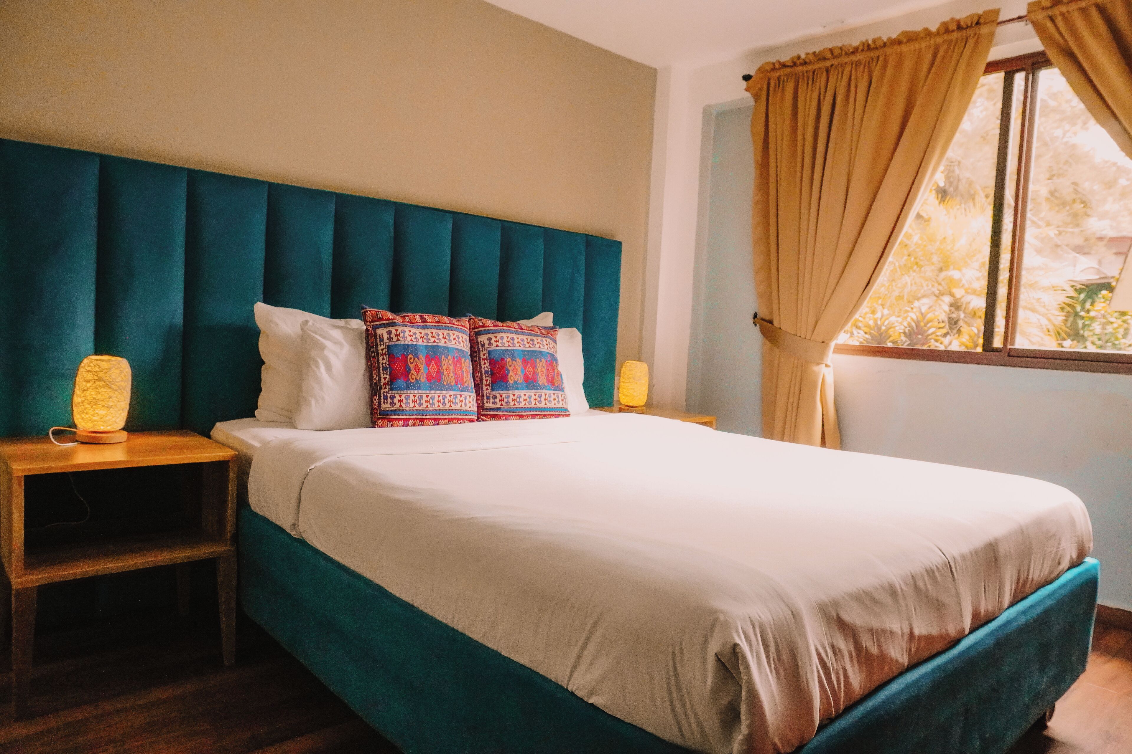 Un lit avec une tête de lit de couleur turquoise, une literie blanche et des coussins à motifs. Il y a une table de chevet avec une lampe et une petite étagère.