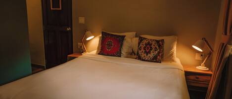 Una cama bien hecha con sábanas blancas y almohadas decorativas, dos lámparas de noche y una puerta de madera.