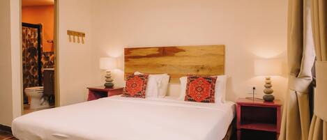 Une chambre d’hôtel avec un lit en bois, deux tables de chevet avec des lampes, une tête de lit en bois et une vue sur la salle de bain par une porte ouverte.