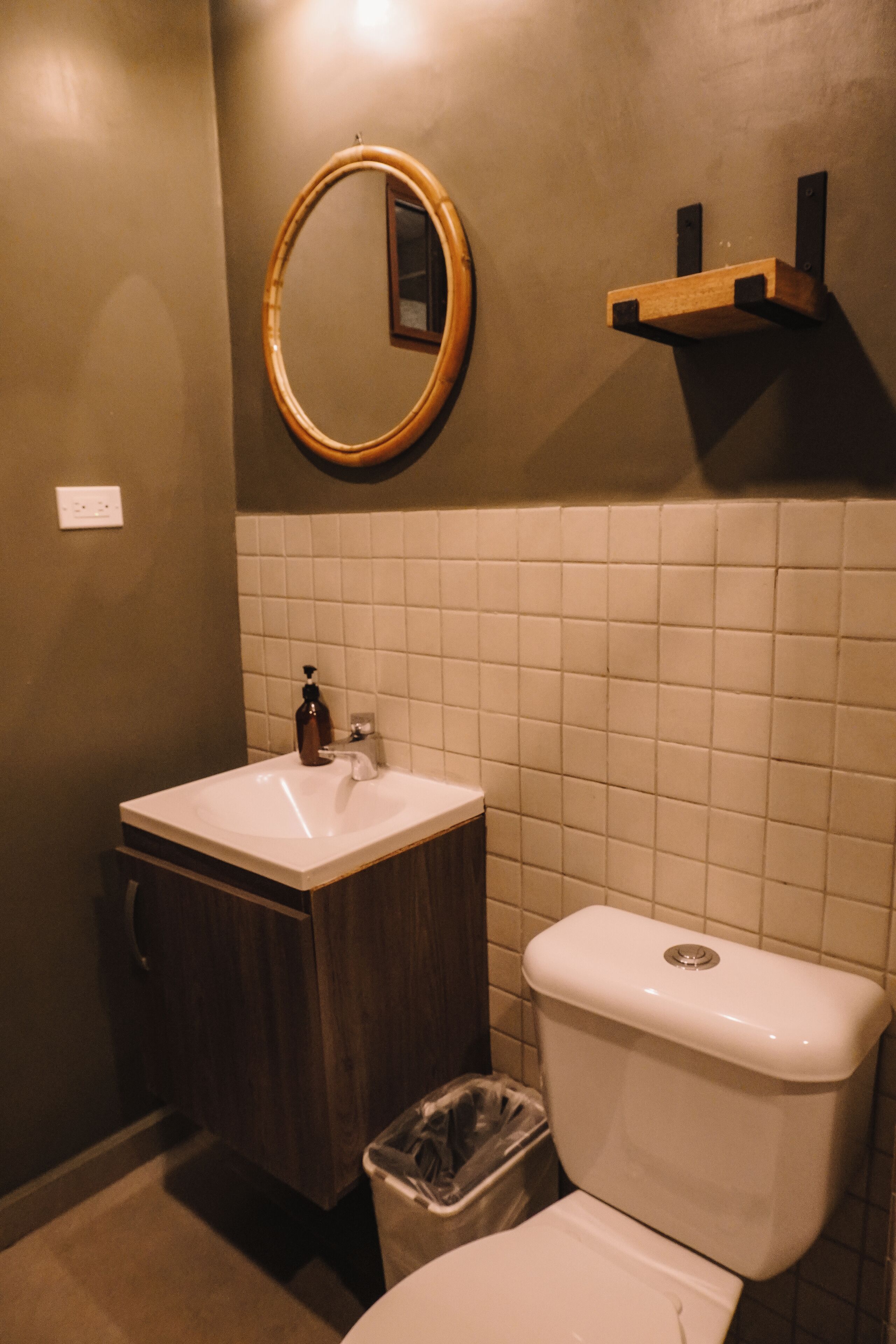 A bathroom with a white sink, wooden vanity, a toilet, a trash bin, a mirror, and a shelf.